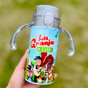 Puede incluir: Un vaso para sorber de pl&aacute;stico transparente con una envoltura azul y verde con el texto "La Granja de Liam" y animales de granja de dibujos animados. La taza tiene una tapa transparente, dos asas y un cierre a presi&oacute;n.