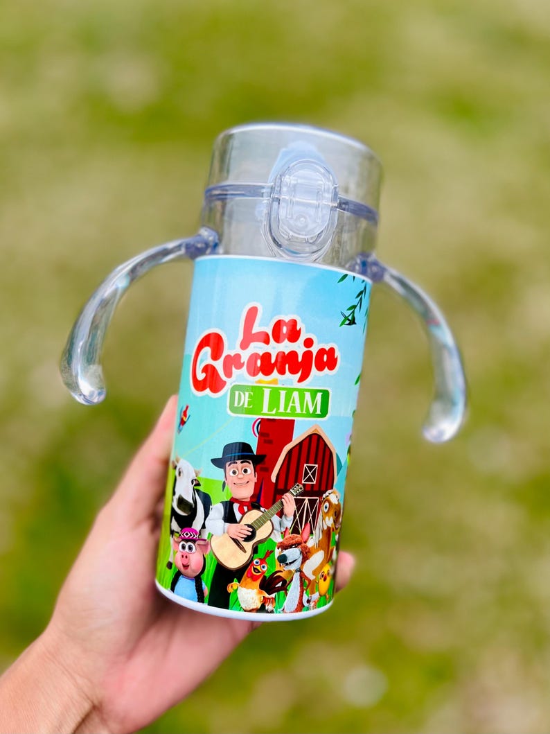 Puede incluir: Un vaso para sorber de pl&aacute;stico transparente con una envoltura azul y verde con el texto "La Granja de Liam" y animales de granja de dibujos animados. La taza tiene una tapa transparente, dos asas y un cierre a presi&oacute;n.