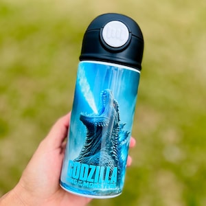 May include: A stainless steel water bottle featuring a Godzilla graphic. The design shows Godzilla with a blue and white background and the text "GODZILLA KING OF THE MONSTERS". The bottle has a black lid with a white button.