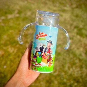 Puede incluir: Vaso para sorber de pl&aacute;stico transparente con una envoltura azul y blanca con dibujos de animales de granja y el texto "La Granja de Zen&oacute;n". El vaso tiene dos asas transparentes y una tapa transparente.