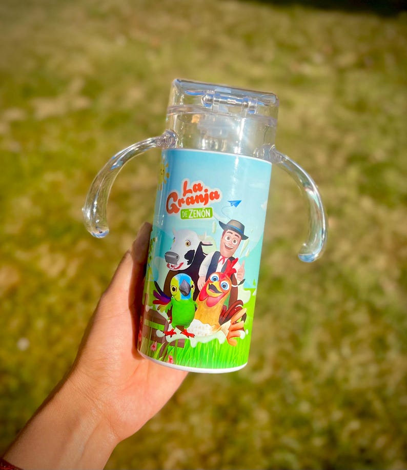 Puede incluir: Vaso para sorber de pl&aacute;stico transparente con una envoltura azul y blanca con dibujos de animales de granja y el texto "La Granja de Zen&oacute;n". El vaso tiene dos asas transparentes y una tapa transparente.