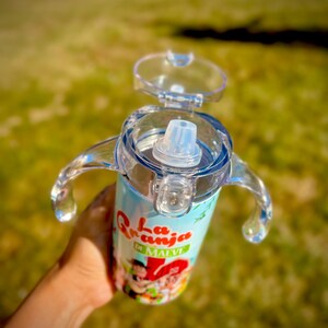 Puede incluir: Un vaso para sorber de color azul claro y blanco con tapa y asas transparentes. El vaso presenta un dise&ntilde;o de dibujos animados colorido con el texto "La Granja de Malve". El vaso est&aacute; dise&ntilde;ado para ni&ntilde;os peque&ntilde;os y ni&ntilde;os.