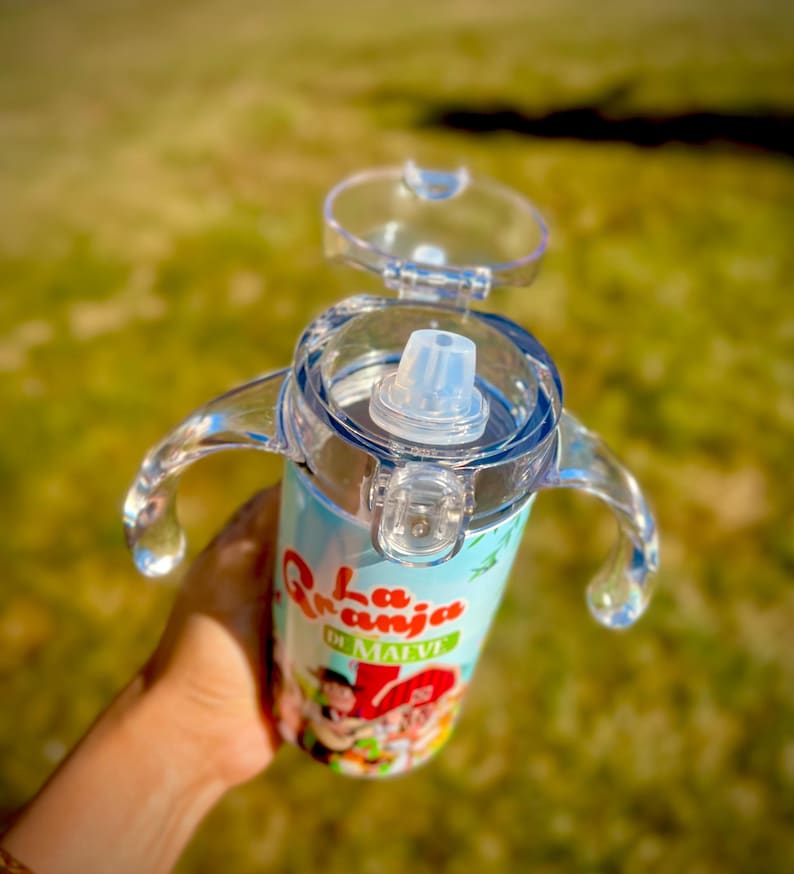 Puede incluir: Un vaso para sorber de color azul claro y blanco con tapa y asas transparentes. El vaso presenta un dise&ntilde;o de dibujos animados colorido con el texto "La Granja de Malve". El vaso est&aacute; dise&ntilde;ado para ni&ntilde;os peque&ntilde;os y ni&ntilde;os.