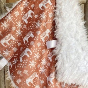 May include: A soft, plush baby blanket with a white faux fur border. The blanket features a rust-colored background with a repeating pattern of white elephants and palm trees.
