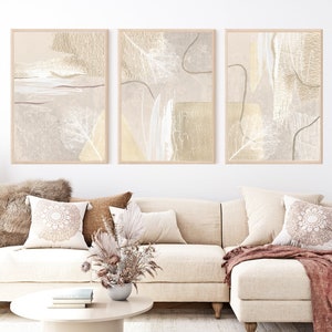 May include: Three abstract art prints in a living room setting. The prints are framed in natural wood and feature a beige and cream color palette with delicate lines and shapes.