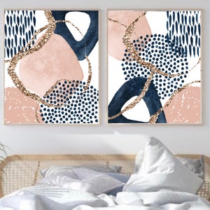 May include: Two framed abstract art prints. Each print features a white background with navy blue, blush pink, and gold glitter accents. The designs include organic shapes, dots, and lines, creating a modern aesthetic. The prints are displayed above a bed.