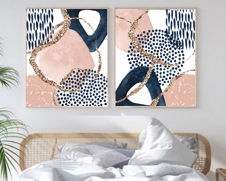 May include: Two framed abstract art prints. Each print features a white background with navy blue, blush pink, and gold glitter accents. The designs include organic shapes, dots, and lines, creating a modern aesthetic. The prints are displayed above a bed.