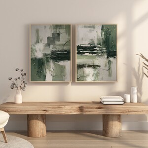 May include: Two abstract paintings in green and white tones are hung on a light beige wall above a wooden bench. The bench has two thick wooden legs and is decorated with a vase of dried pampas grass and a stack of books.