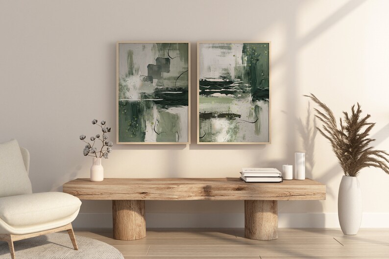 May include: Two abstract paintings in green and white tones are hung on a light beige wall above a wooden bench. The bench has two thick wooden legs and is decorated with a vase of dried pampas grass and a stack of books.