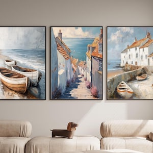 May include: A triptych of framed coastal paintings. The artwork depicts scenes of boats on a beach, a seaside village street, and a coastal town with boats. The paintings feature a muted color palette of blues, whites, and browns.