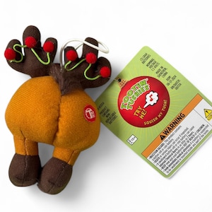 May include: A plush reindeer toy with brown antlers adorned with red pom-poms and green accents. The reindeer has an orange body, brown legs, and a tag that reads "Tootin' Tushies" with a warning about batteries. The toy is designed to be squished.