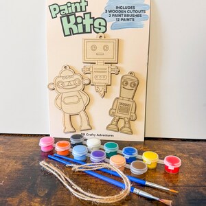 May include: A paint kit featuring three wooden robot cutouts, 12 paint pots in various colors, and two paint brushes. The kit includes a card with the words "Paint Kits" and the text "Includes 3 Wooden Cutouts, 2 Paint Brushes, 12 Paints."