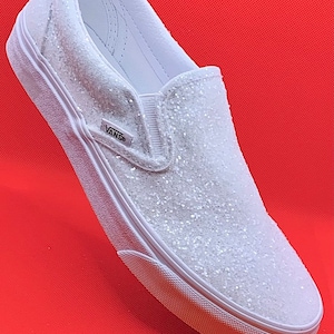 May include: White slip-on Vans sneakers with a sparkly white glitter finish. The Vans logo is visible on the side of the shoe.