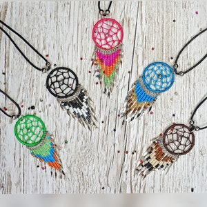 May include: Five dreamcatchers with beaded fringe hanging from black cords. The dreamcatchers are made of macrame and have different coloured beads. The dreamcatchers are on a white wooden background.