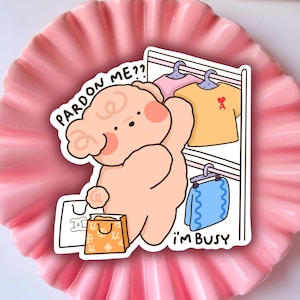 May include: A pink cartoon bear with a shopping bag and the text "Pardon me? I'm busy" is holding a hanger with a yellow shirt and a blue towel on it.