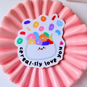 Puede incluir: Una pegatina blanca con una ilustración de dibujos animados de un tazón de cereal con piezas de cereal de colores que se derraman. El texto "cereal-sly love you" está escrito alrededor del tazón.