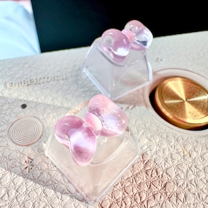 May include: Two clear plastic keycaps with pink bow-shaped keycap toppers. The keycaps are on a light brown leather surface.
