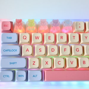 Pode incluir: Um teclado branco com teclas azul pastel e rosa. O teclado tem uma barra de espa&ccedil;o rosa e uma linha de teclas em forma de ursinhos de goma.