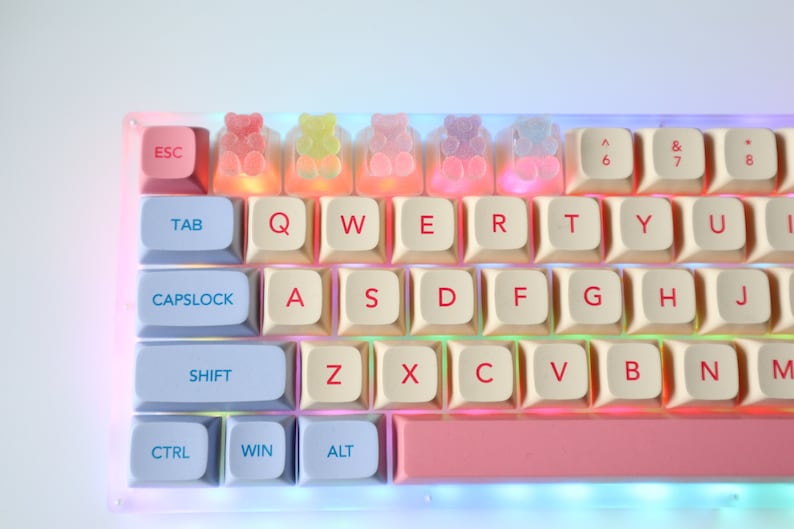 Pode incluir: Um teclado branco com teclas azul pastel e rosa. O teclado tem uma barra de espa&ccedil;o rosa e uma linha de teclas em forma de ursinhos de goma.