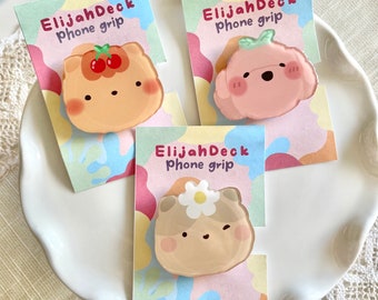 May include: Three phone grips with cute cartoon animal designs. The first grip is orange with a cherry on top, the second is pink with a green leaf, and the third is brown with a white flower. Each grip has the text "ElijahDeck phone grip" on the packaging.