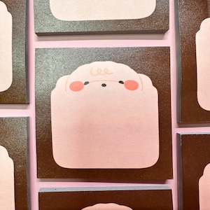 May include: A collection of square sticky notes with a light pink sheep design on a brown background. The sheep has pink cheeks and a curly line for the hair. The background is a light pink color.