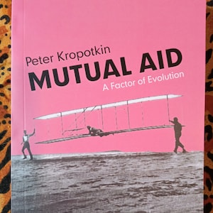 May include: A book titled "MUTUAL AID A Factor of Evolution" by Peter Kropotkin. The cover is pink with black text and a black and white image of a biplane with people. The background is a pink sky.