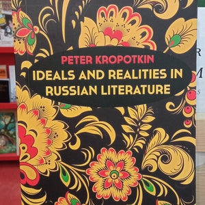May include: A book titled "Ideals and Realities in Russian Literature" by Peter Kropotkin. The cover features a black background with a floral design in yellow, red, and green. The book is displayed on a red surface.