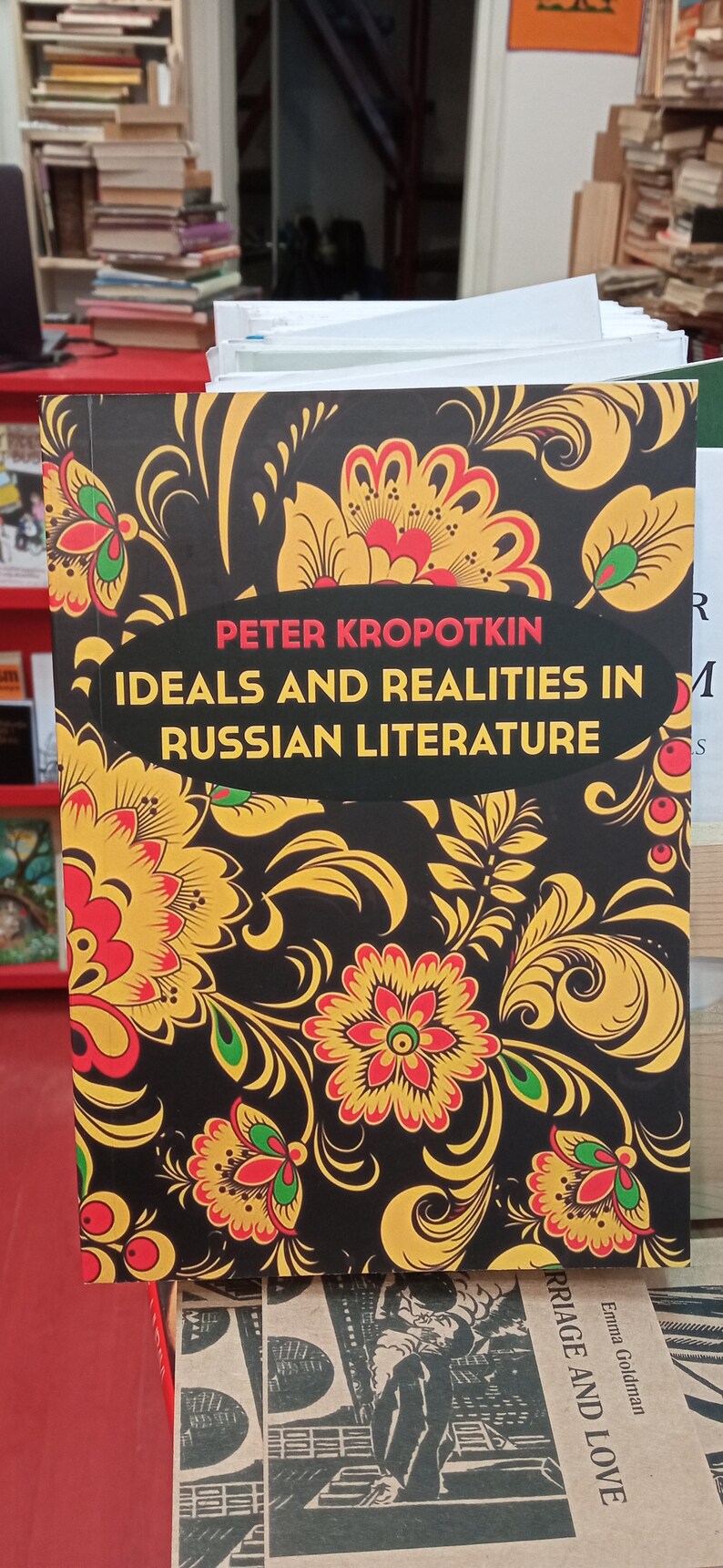May include: A book titled "Ideals and Realities in Russian Literature" by Peter Kropotkin. The cover features a black background with a floral design in yellow, red, and green. The book is displayed on a red surface.