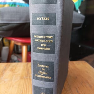 以下が含まれることがあります： 金色の文字が書かれたグレーのハードカバーの本。本の背表紙には「MYŠKIS」というタイトルと「INTRODUCTORY MATHEMATICS FOR ENGINEERS」と「Lectures in Higher Mathematics」というテキストが書かれています。本は木製の表面にあります。