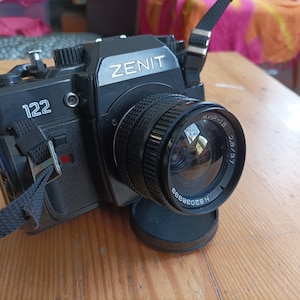 May include: Black Zenit 122 film camera with a black lens and lens cap. The camera has a black strap and the text "ZENIT" and "122" are visible. The lens has the text "MИP-1B 2.8/37".