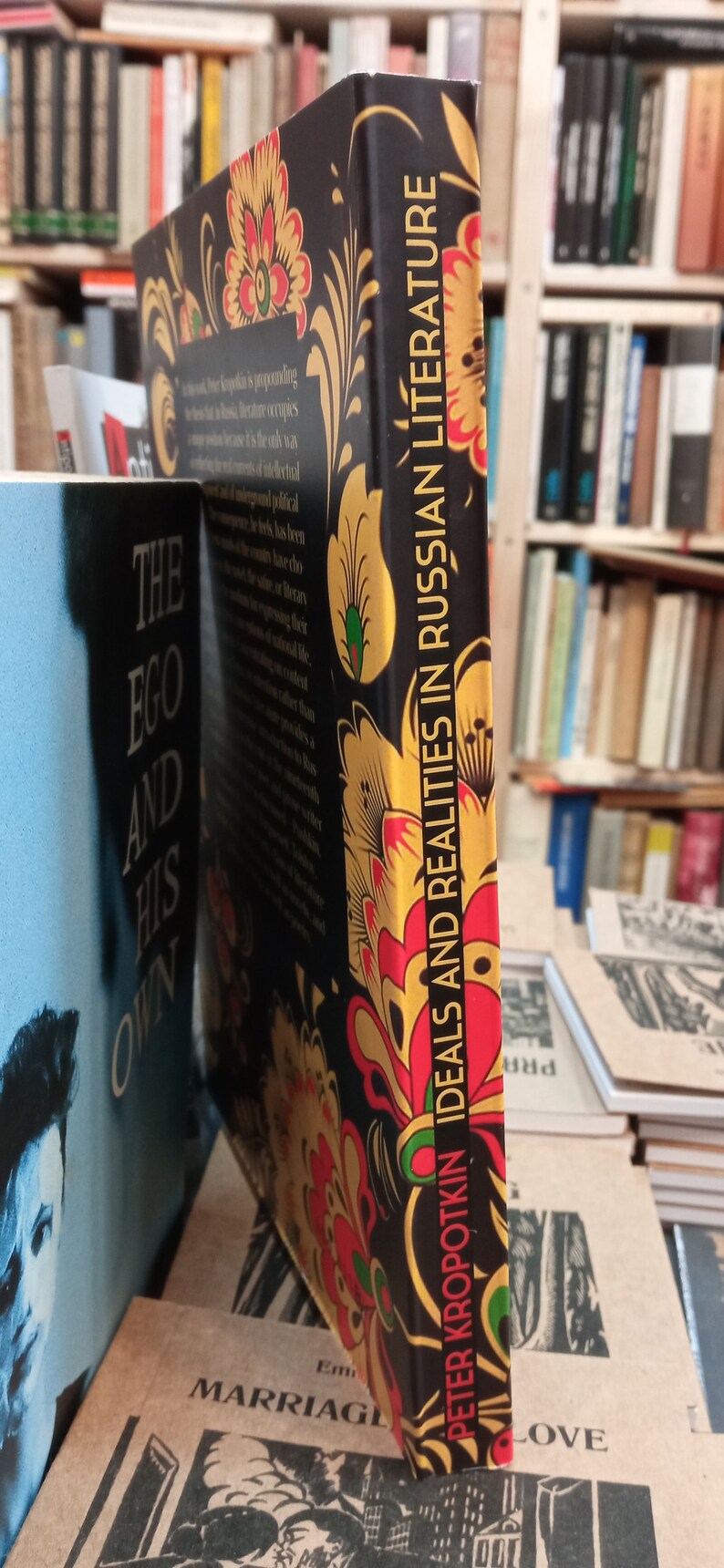 May include: A hardcover book titled "Ideals and Realities in Russian Literature" by Peter Kropotkin. The book has a black cover with a floral design in red, yellow, and gold. The spine displays the title vertically.