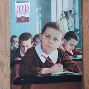 Puede incluir: Portada de revista vintage que presenta una escena de aula con niños con uniformes marrones y cuellos blancos. La portada incluye texto en ruso y el año 1949. La revista está sobre una superficie de madera.