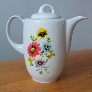 May include: White ceramic teapot with a lid and a curved handle. The teapot is decorated with a floral design featuring red, yellow, and blue flowers with green leaves. The teapot is on a wooden surface.