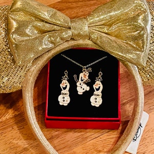May include: Gold glitter Minnie Mouse ears with a gold bow. A red box with a black interior contains a necklace and earrings featuring Olaf from Frozen. The box has a Disney label.