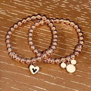 May include: Two rose gold beaded bracelets with charms. One charm is a heart with a Mickey Mouse silhouette inside. The other charm is a white Mickey Mouse head.