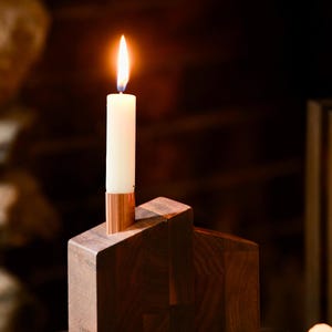 May include: A lit white candle in a copper holder sits atop a geometric wooden candle holder. The candle holder is made of dark brown wood with a copper accent. The flame is bright and the background is blurred.