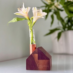 May include: A small wooden house-shaped vase with a copper tube holding two white and yellow flowers. The vase has a geometric design with a dark purple and brown color scheme. The text "Hauskaa Verkkari" is visible on the front.