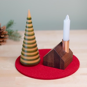 Puede incluir: Un portavelas de madera con forma de casa con un portavelas de cobre y una vela blanca. Un árbol de Navidad de madera con rayas verdes y marrones se encuentra sobre una alfombra de fieltro roja.