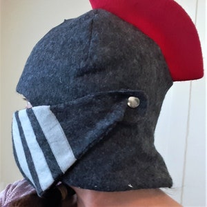 May include: A dark gray helmet with a red crest. The helmet has a visor with three white stripes and a silver button closure. The headwear is likely for costume or historical reenactment.