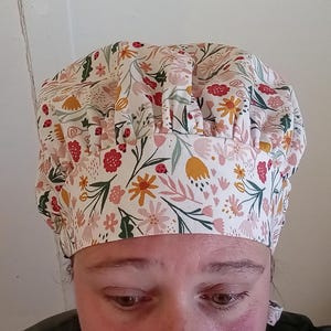 Op de afbeelding: Een witte scrubmuts van stof met een bloemenprint met roze, gele en rode bloemen. De muts heeft een geplooide bovenkant en is ontworpen om in een medische omgeving te worden gedragen.