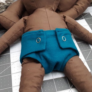 May include: A brown plush toy with a teal diaper. The diaper has two silver snaps and a pocket on each side.