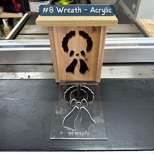 May include: A wooden box with a cut-out design of a wreath and bells. The box is sitting on a black surface next to a clear acrylic stencil with the same design. The stencil has the text "#8 Wreath" printed on it.