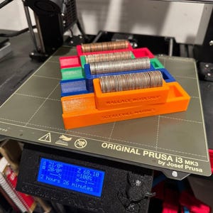 May include: A 3D printed coin holder with four compartments, each holding a stack of coins. The holder is printed in orange, blue, green, and red plastic. The holder is labeled "WALLACE WORKSHOP" and "SCT KEEVILA MARINES". The holder is sitting on a black and gray 3D printer bed. The printer screen shows the progress of the print job.