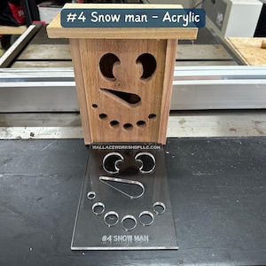 May include: A wooden snowman cutout with a face and buttons. The cutout is on a clear acrylic sheet with the text "#4 SNOW MAN" printed on it.