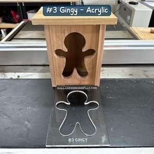 May include: A wooden box with a gingerbread man cutout. The box is sitting on a black surface. A clear acrylic sheet with a gingerbread man cutout is in front of the box. The acrylic sheet has the text "#3 GINGY" printed on it.
