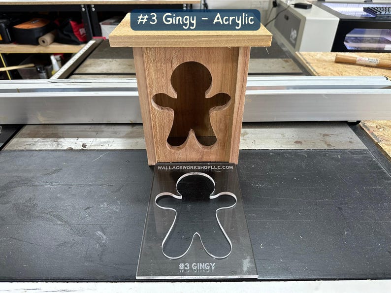 May include: A wooden box with a gingerbread man cutout. The box is sitting on a black surface. A clear acrylic sheet with a gingerbread man cutout is in front of the box. The acrylic sheet has the text "#3 GINGY" printed on it.
