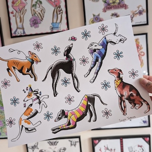 May include: A white art print featuring six greyhound dogs in various poses and outfits, including sweaters and bandanas, surrounded by small blue and pink flower illustrations. The bottom right corner contains the handwritten text Snips Tattoos.