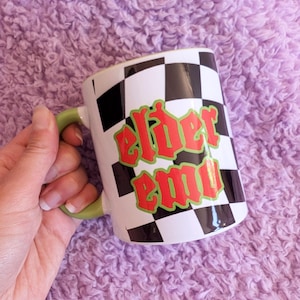 May include: White ceramic mug with a black and white checkered pattern. The mug has green and red text that says "elder emo".