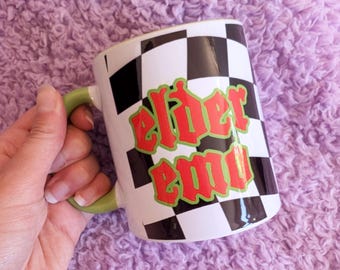 May include: White ceramic mug with a black and white checkered pattern. The mug has green and red text that says "elder emo".