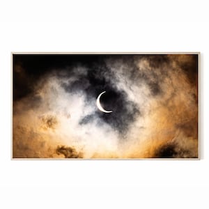 Pode incluir: Impressão emoldurada com um eclipse solar. A imagem mostra um sol em forma de crescente rodeado por nuvens escuras e rodopiantes, com tons de laranja e amarelo. A obra de arte é emoldurada em uma moldura de madeira clara.